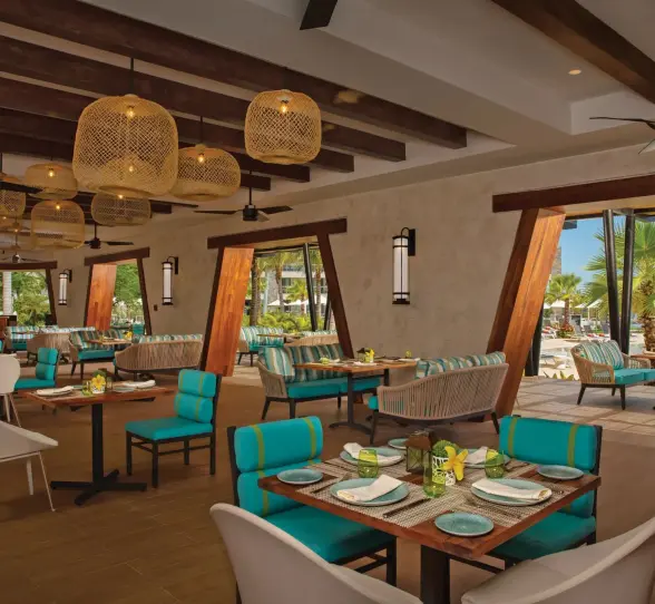 Stylish open-air restaurant with turquoise seating, wooden beams, and poolside views surrounded by palm trees.