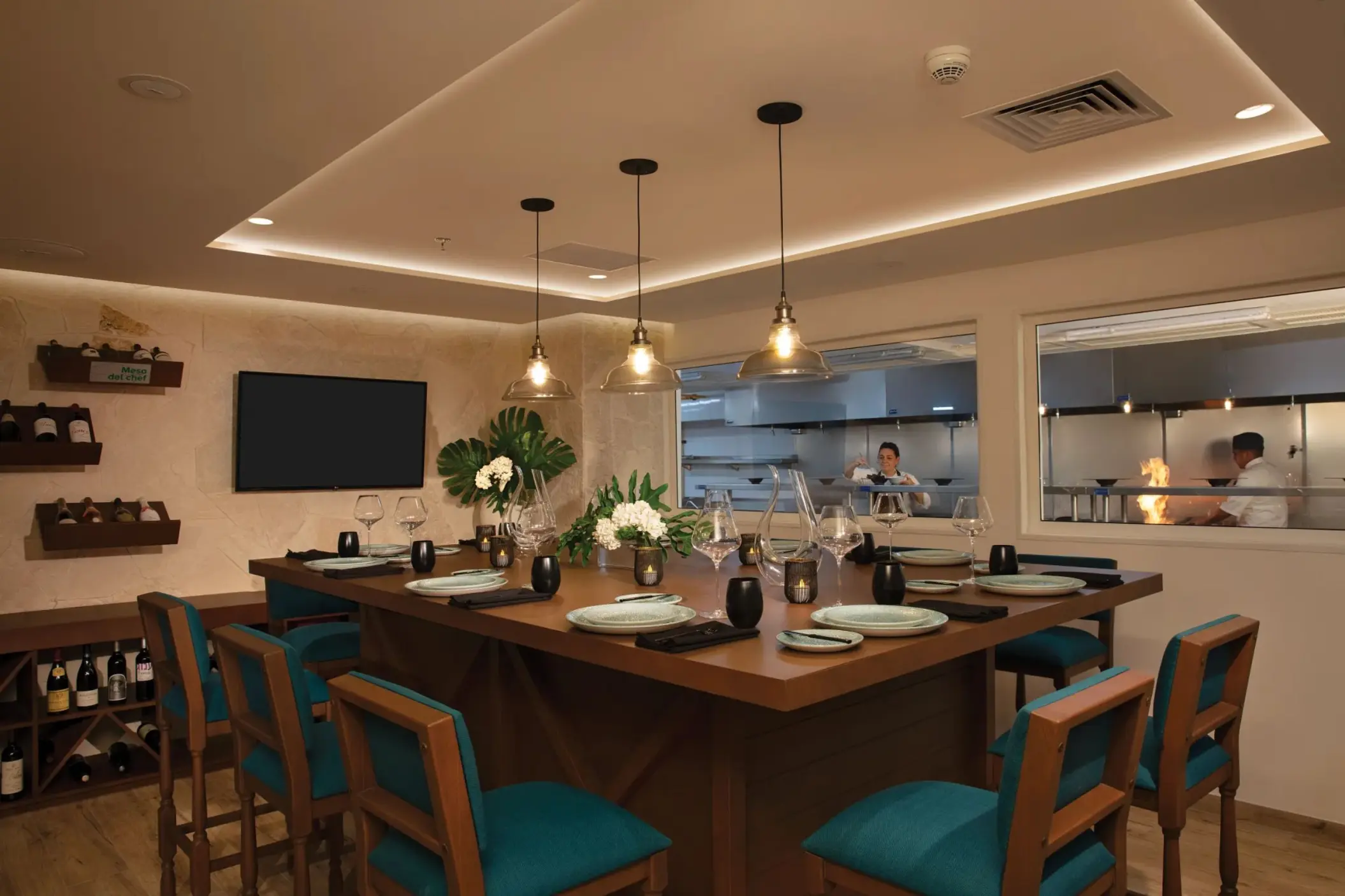 Private chef’s table dining room with elegant table settings and open kitchen view where chefs are preparing meals.