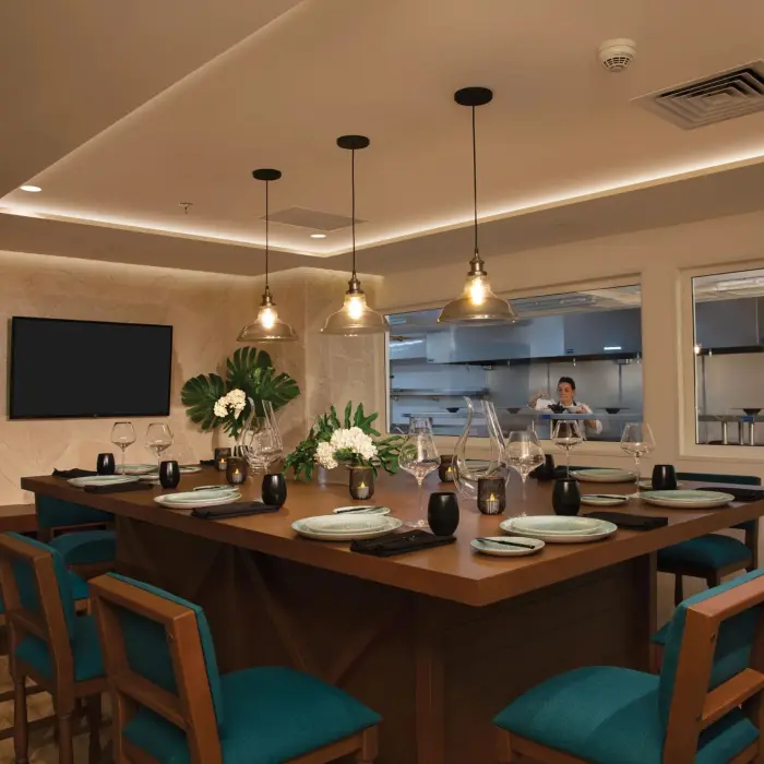 Private chef’s table dining room with elegant table settings and open kitchen view where chefs are preparing meals.