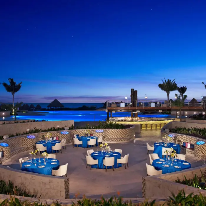 Poolside gala dinner at night with ocean backdrop, round blue-covered tables, and romantic lighting.