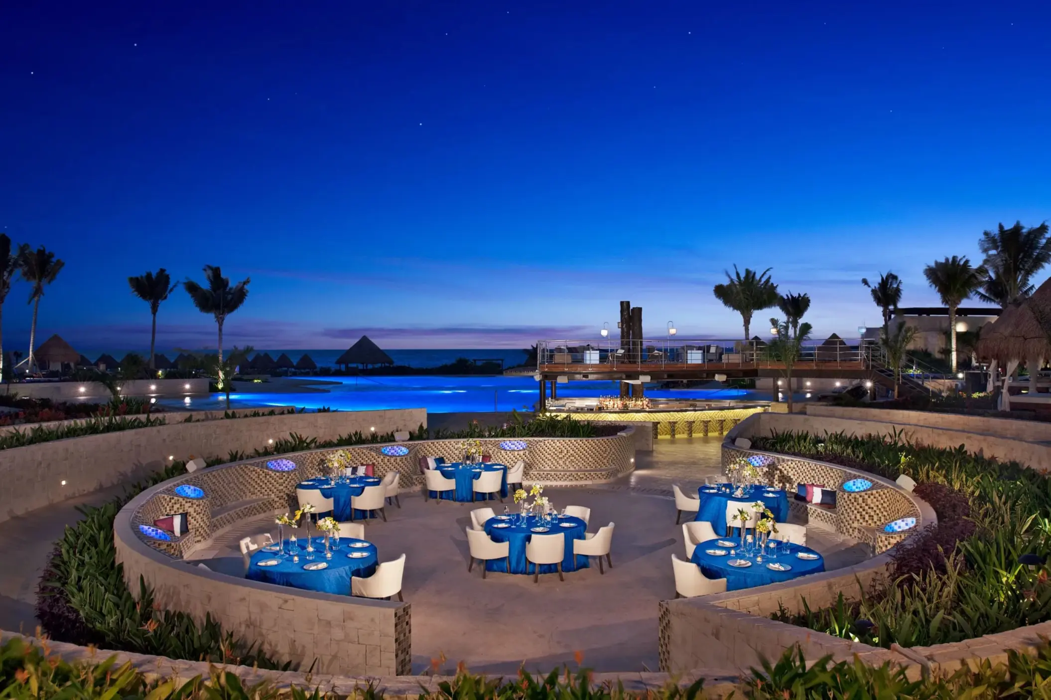 Poolside gala dinner at night with ocean backdrop, round blue-covered tables, and romantic lighting.