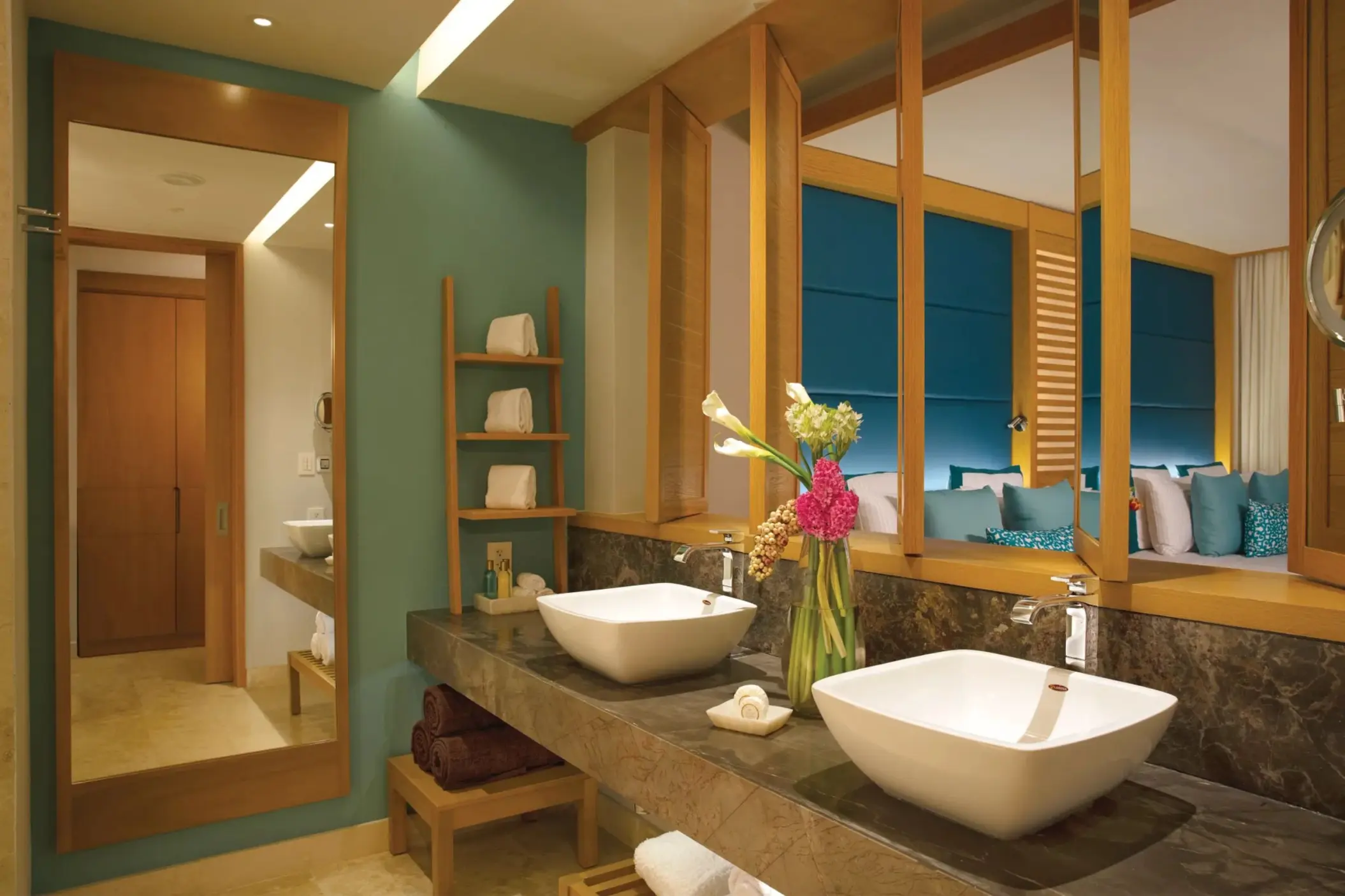 Modern bathroom with dual sinks, marble counter, and peekaboo view into suite with teal headboard.