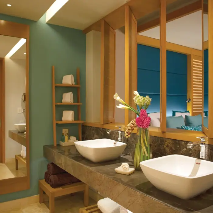 Modern bathroom with dual sinks, marble counter, and peekaboo view into suite with teal headboard.