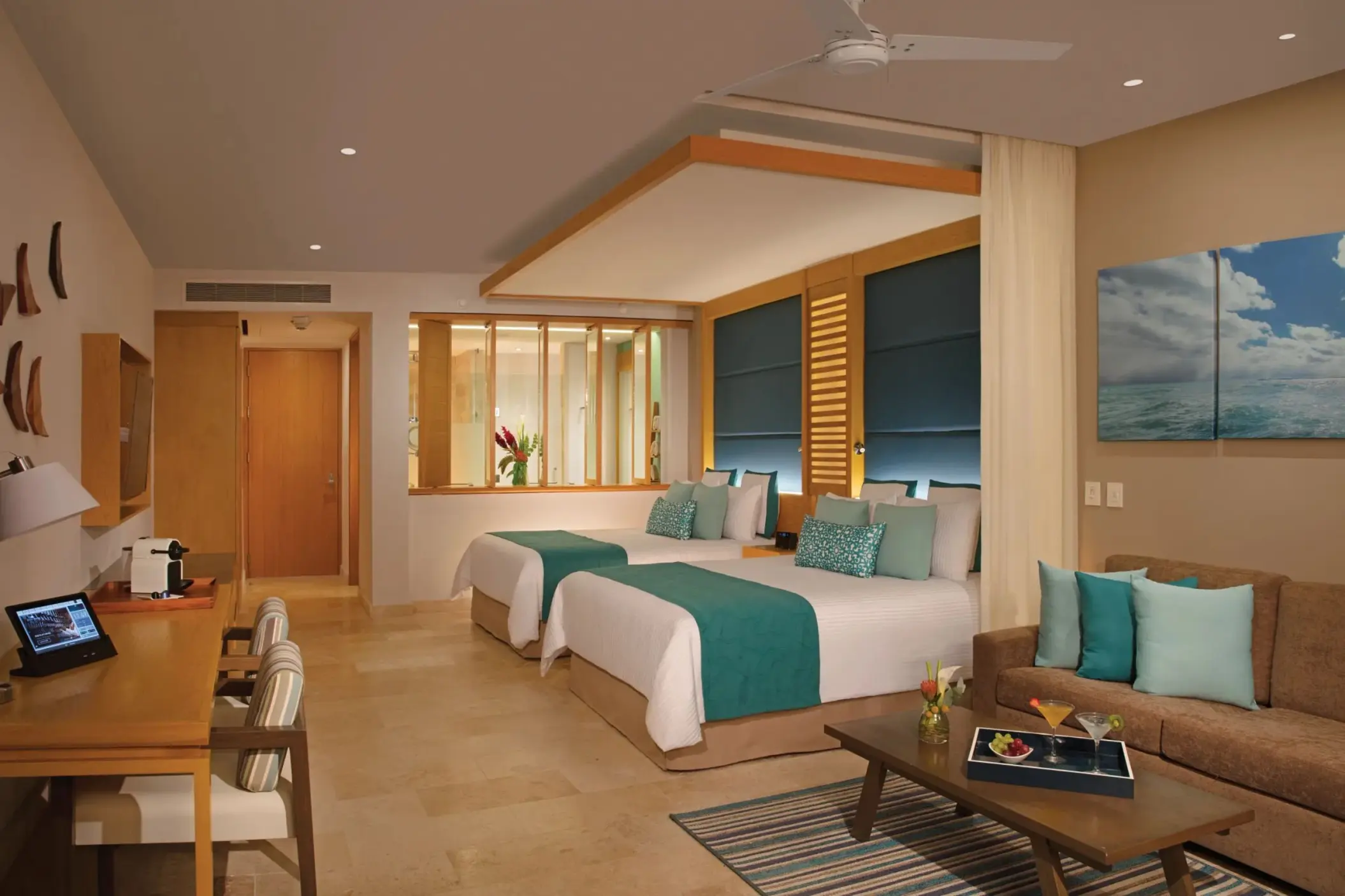 Modern junior suite with two beds, plush bench seating, and open bathroom layout with dual sinks.