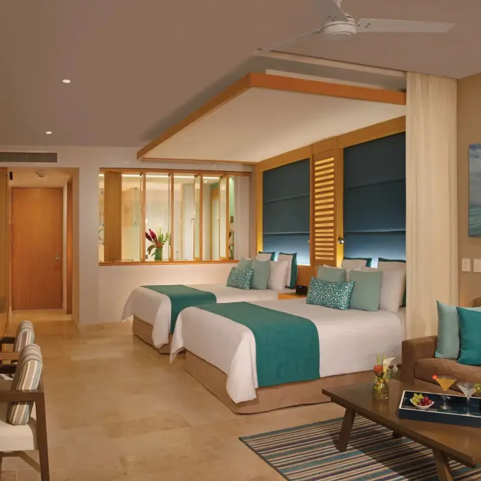 Modern junior suite with two beds, plush bench seating, and open bathroom layout with dual sinks.