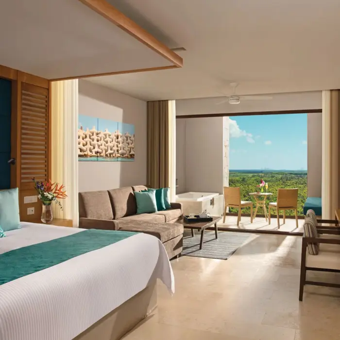 Spacious junior suite with king bed, sofa area, and balcony overlooking lush green tropical scenery.