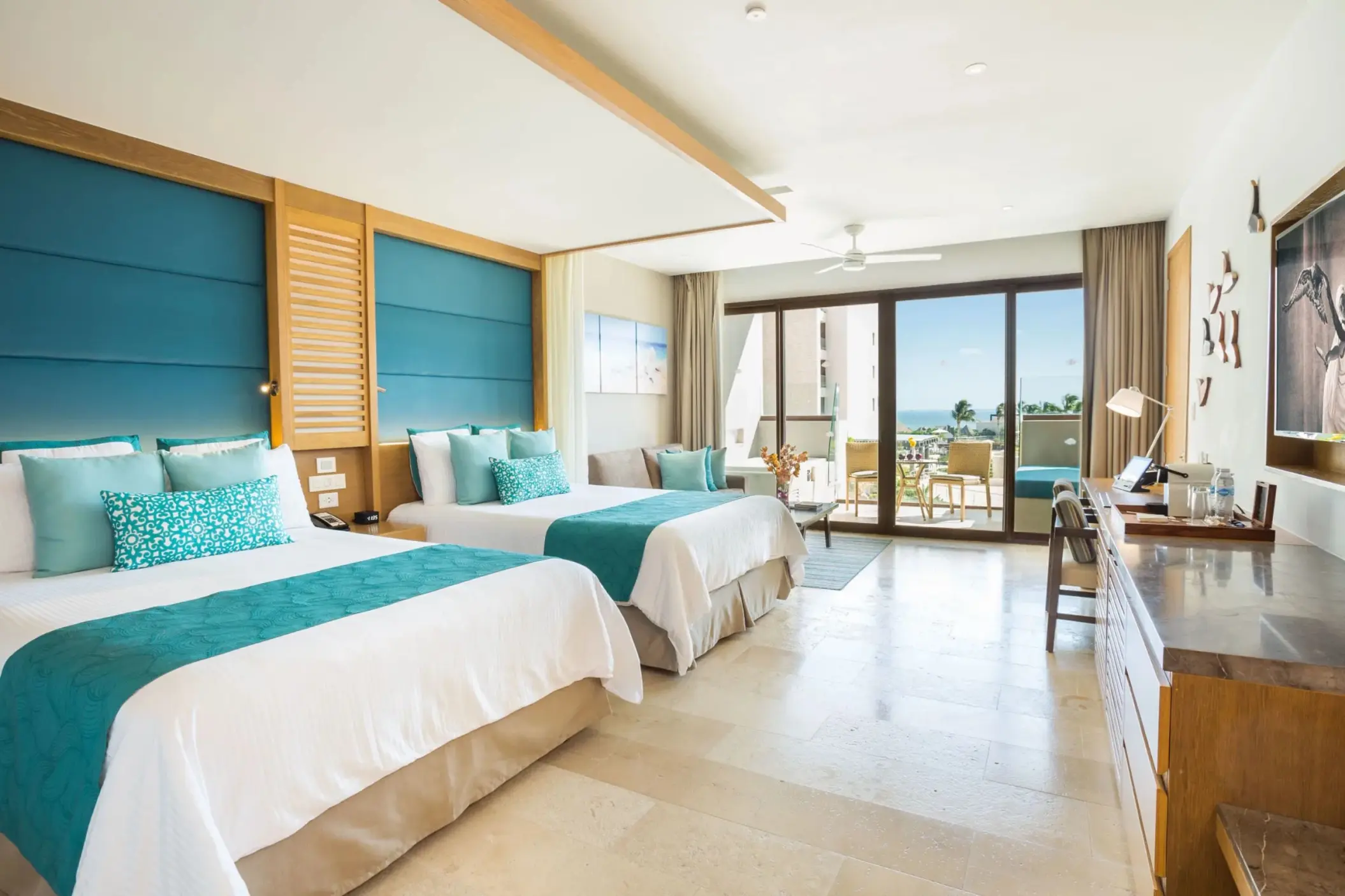 Bright junior suite with double beds, teal accents, and ocean view balcony at Dreams Playa Mujeres.
