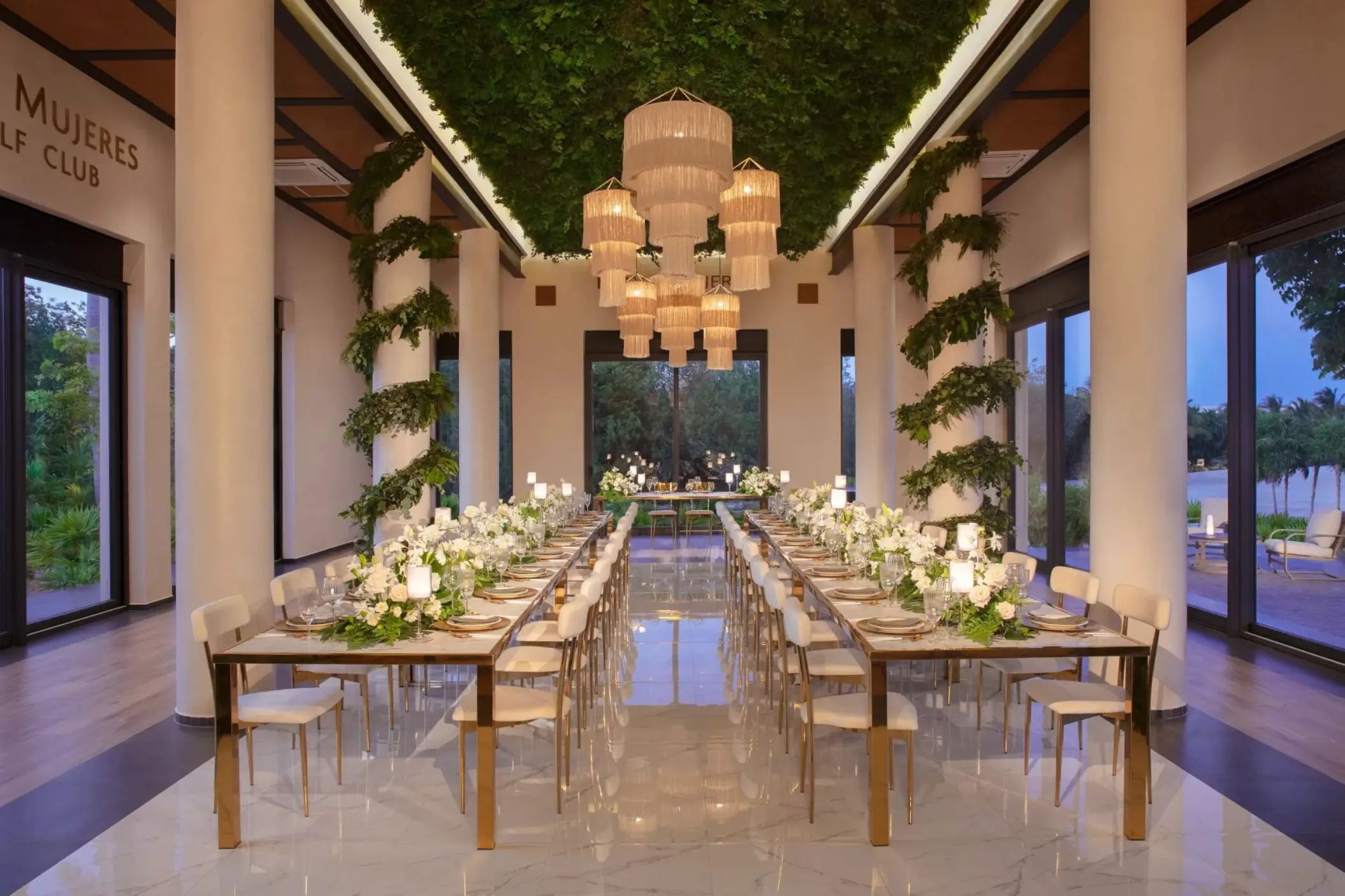 Elegant wedding reception setup in a bright ballroom with lush greenery, chandeliers, and mirrored tables.