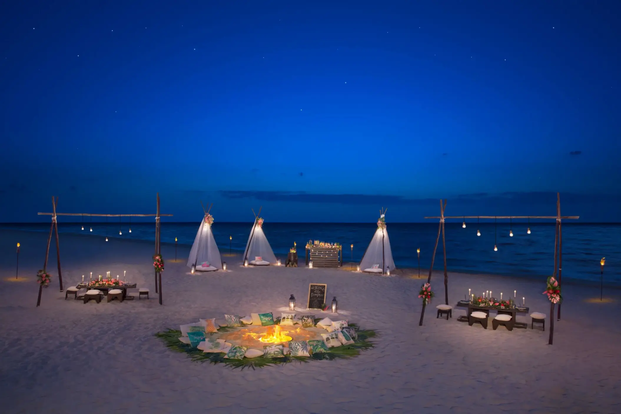 Magical beachfront event setup with teepee tents and firepit under stars.