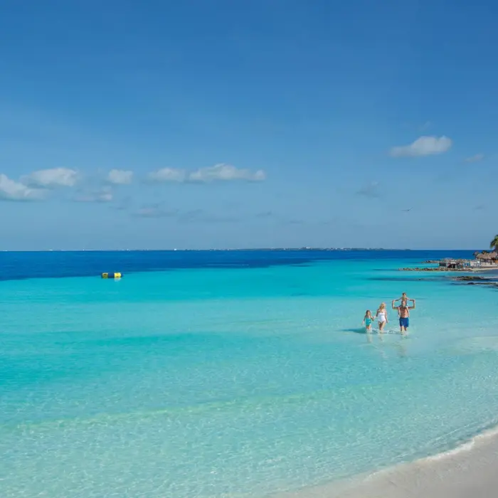 Family walking through crystal-clear shallow waters of resort beach area.