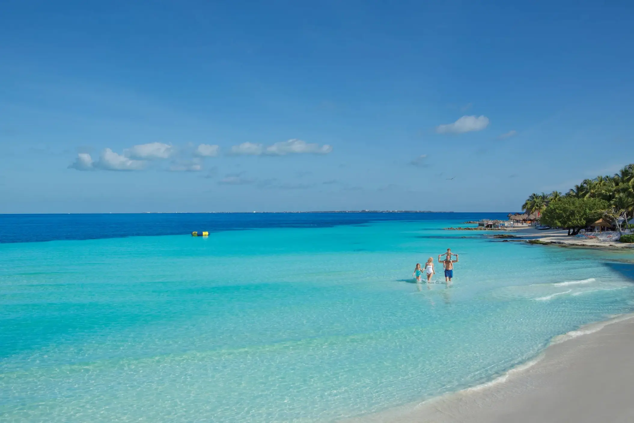 Family walking through crystal-clear shallow waters of resort beach area.
