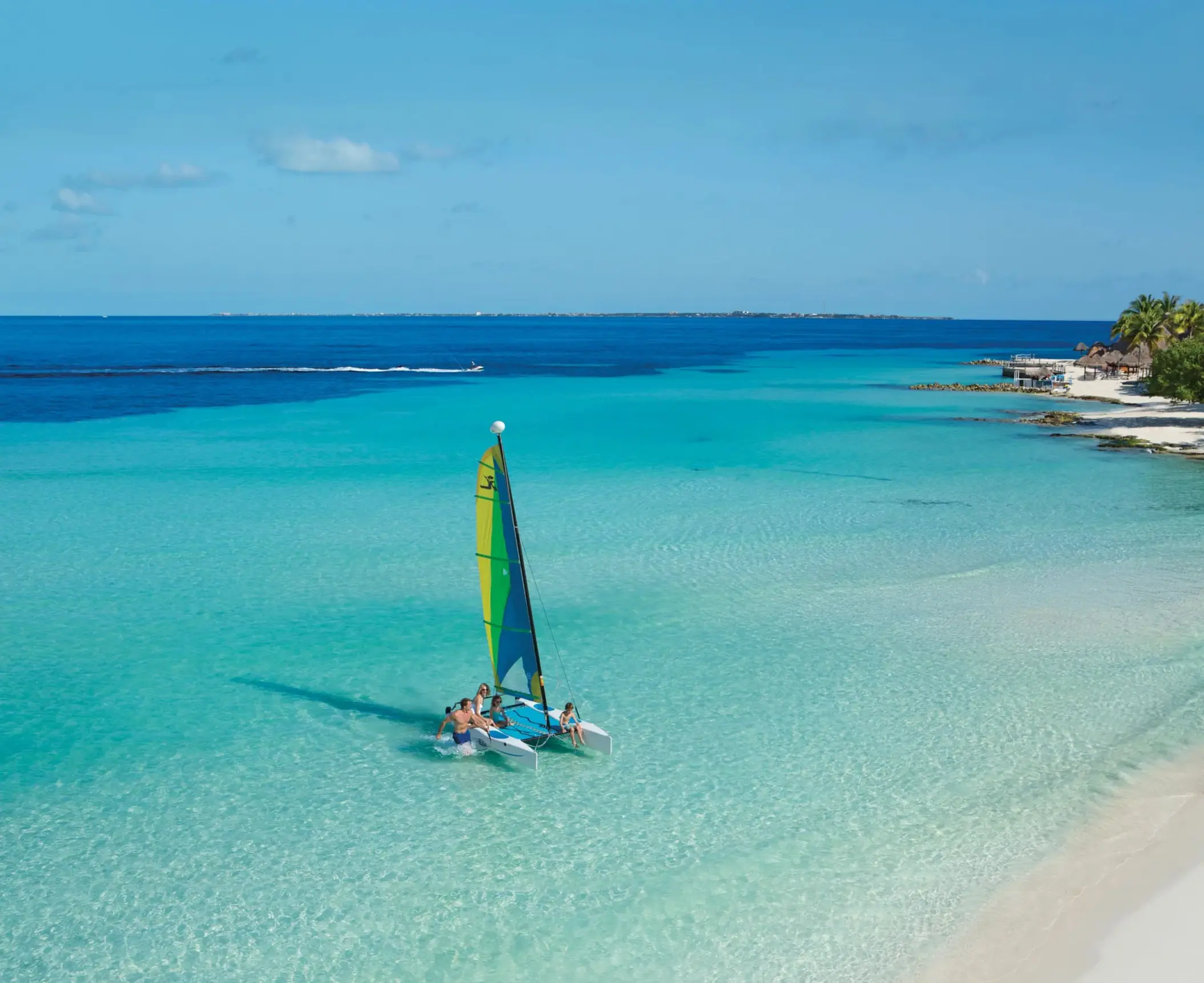 Family on small sailboat in shallow turquoise sea near tropical shoreline.