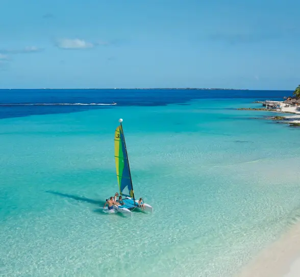 Family on small sailboat in shallow turquoise sea near tropical shoreline.