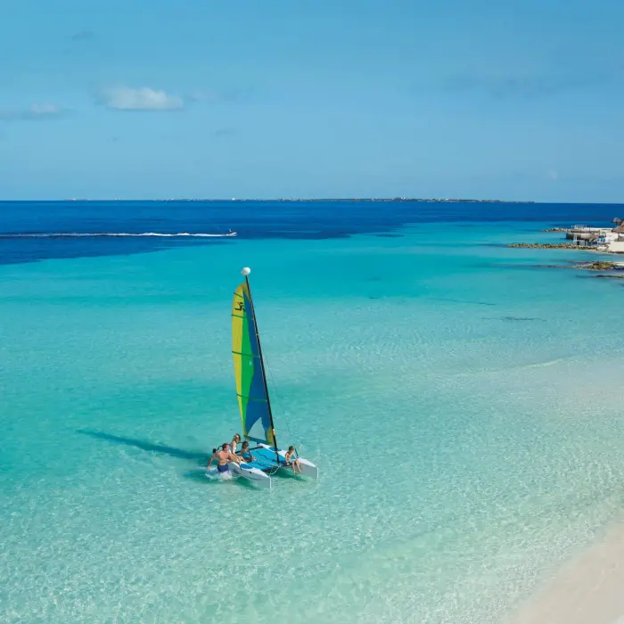 Family on small sailboat in shallow turquoise sea near tropical shoreline.
