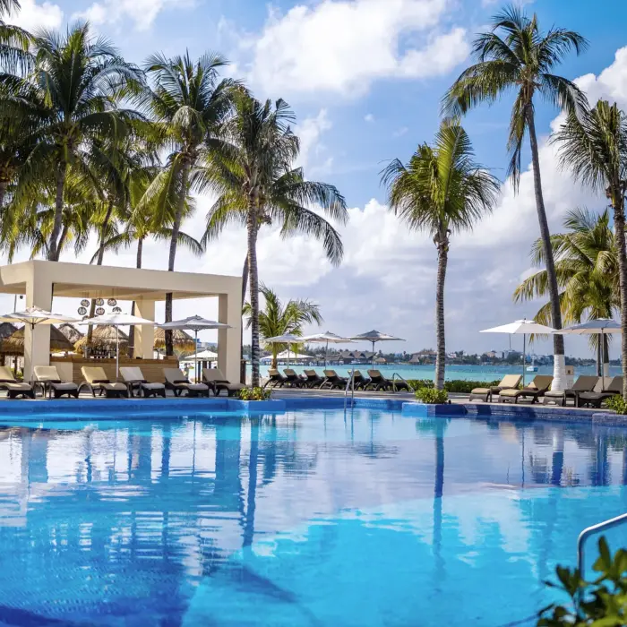Infinity-edge pool with ocean view surrounded by palm trees, loungers, and a modern poolside bar pavilion.