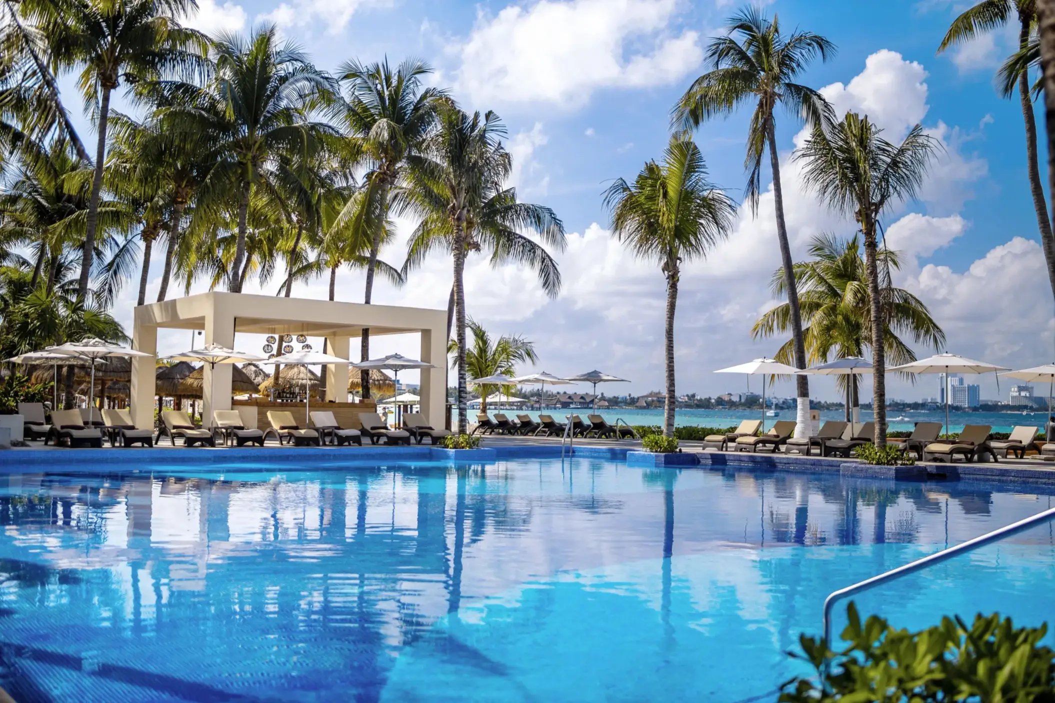Infinity-edge pool with ocean view surrounded by palm trees, loungers, and a modern poolside bar pavilion.