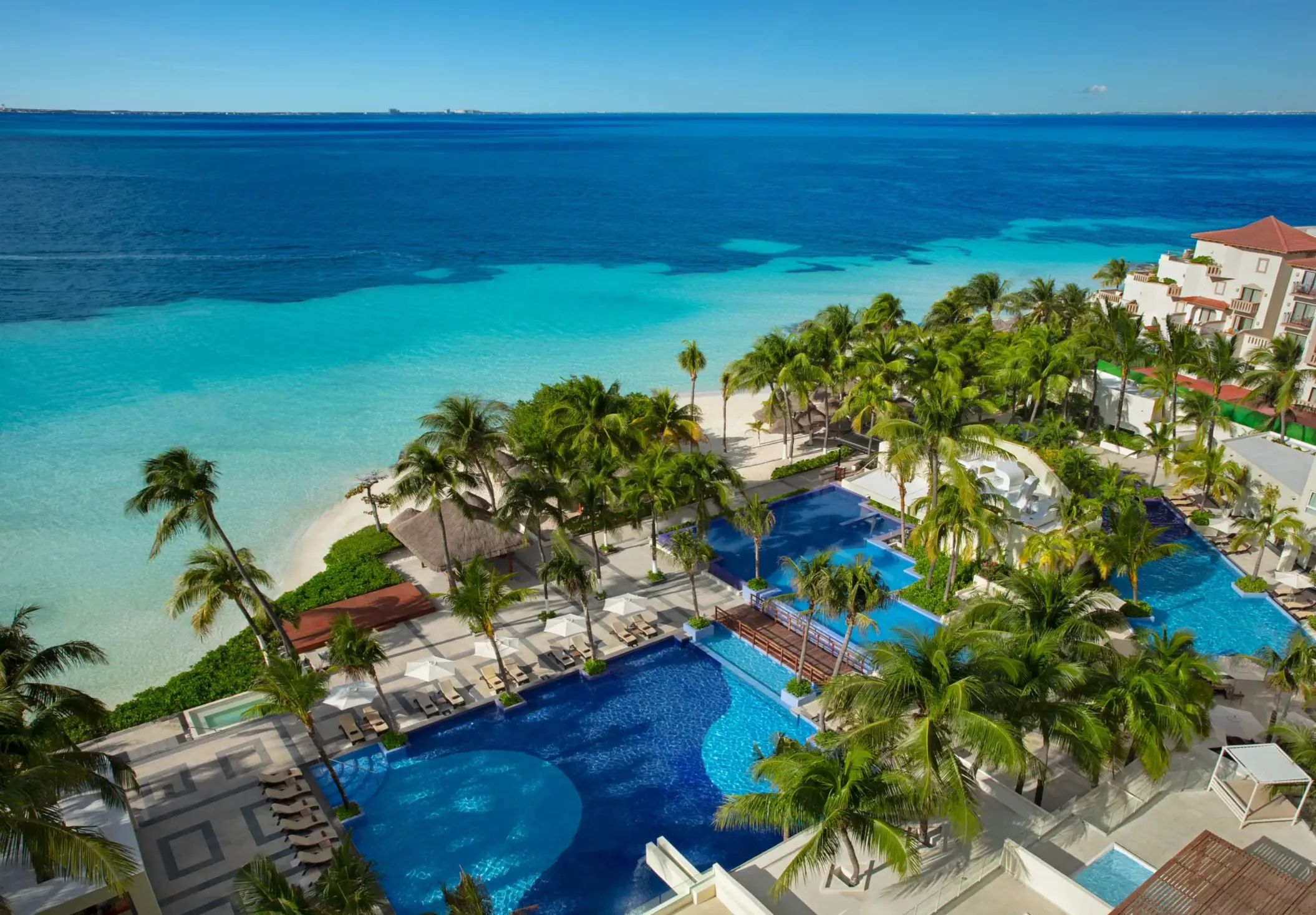 Aerial view of multiple pools lined with palm trees and loungers, steps away from the Caribbean Sea.