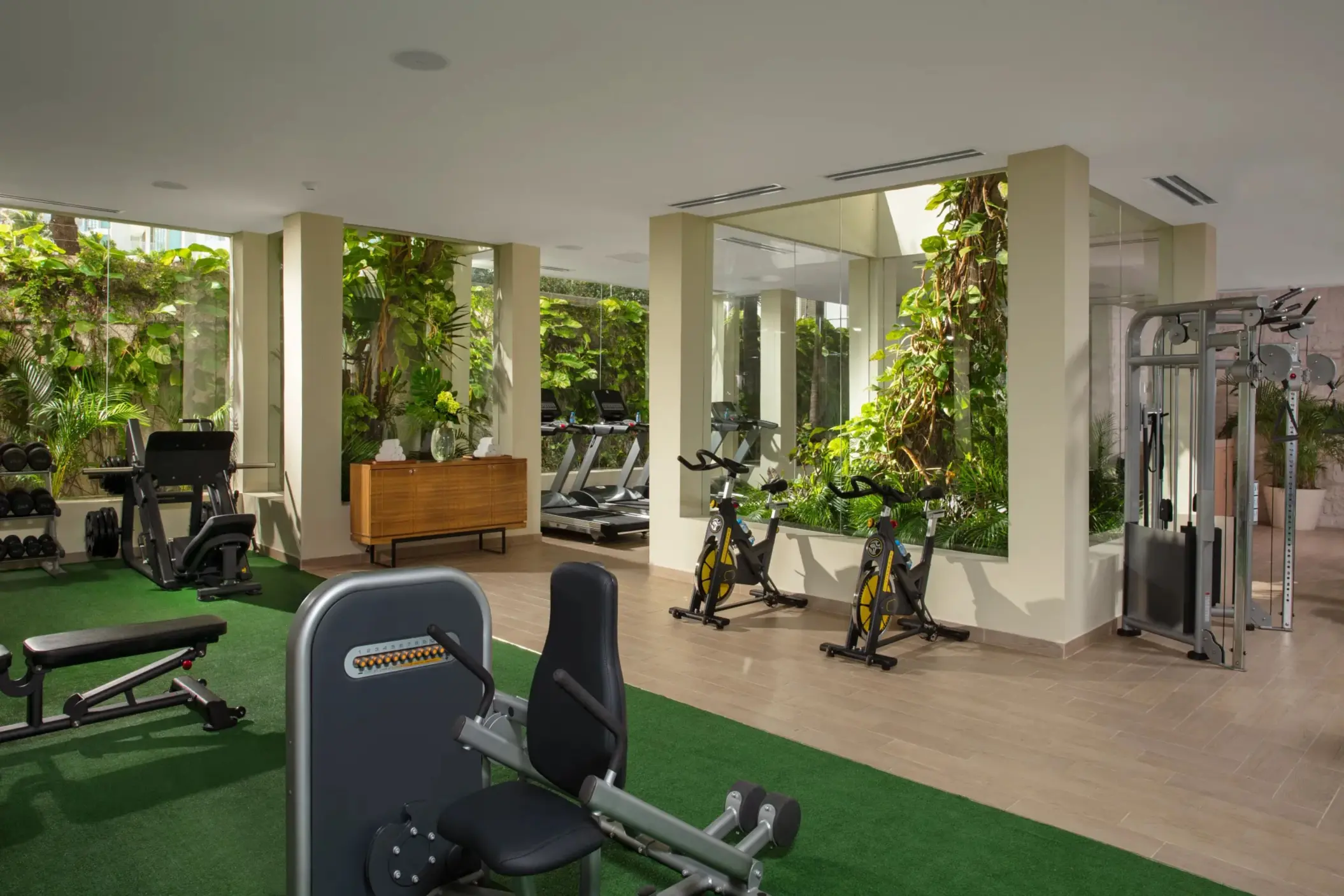 Modern fitness center with cardio machines, weights, and greenery views through floor-to-ceiling windows.