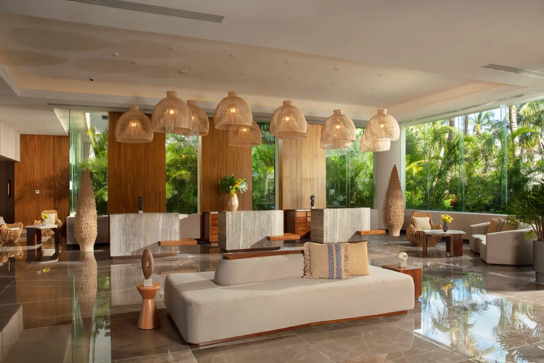 Warm, stylish hotel lobby with woven light fixtures, marble floors, and tropical greenery through tall windows.