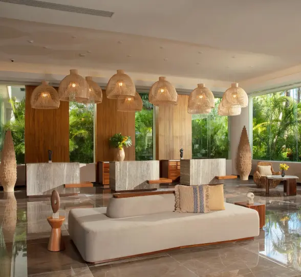 Warm, stylish hotel lobby with woven light fixtures, marble floors, and tropical greenery through tall windows.