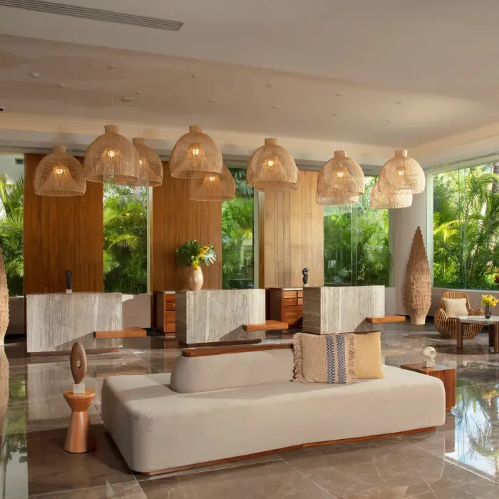 Warm, stylish hotel lobby with woven light fixtures, marble floors, and tropical greenery through tall windows.