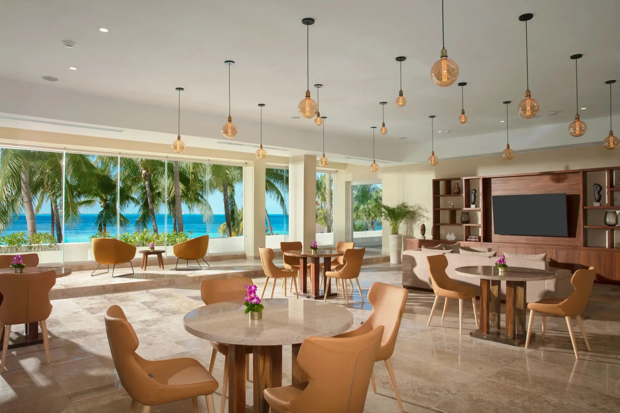 Bright dining lounge with ocean views, modern pendant lights, and tan chairs arranged around round tables.