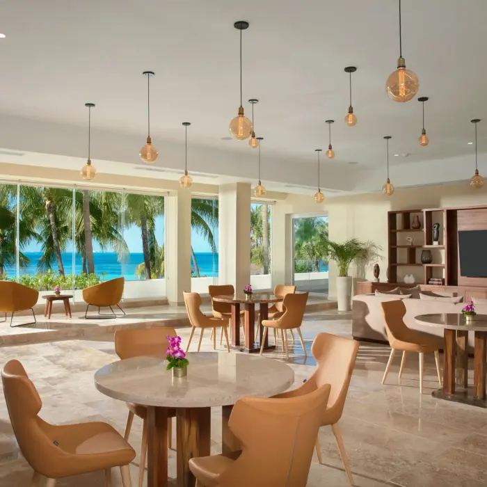 Bright dining lounge with ocean views, modern pendant lights, and tan chairs arranged around round tables.