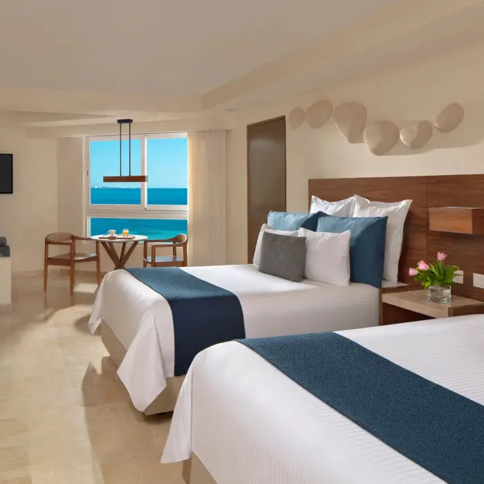 Spacious room with two beds, ocean view dining nook, and soft wood and cream décor throughout the layout.