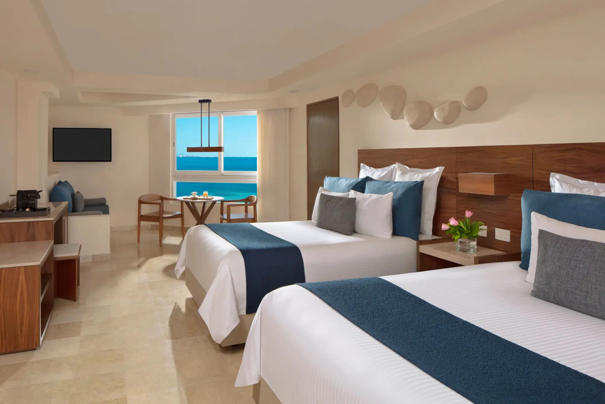 Spacious room with two beds, ocean view dining nook, and soft wood and cream décor throughout the layout.