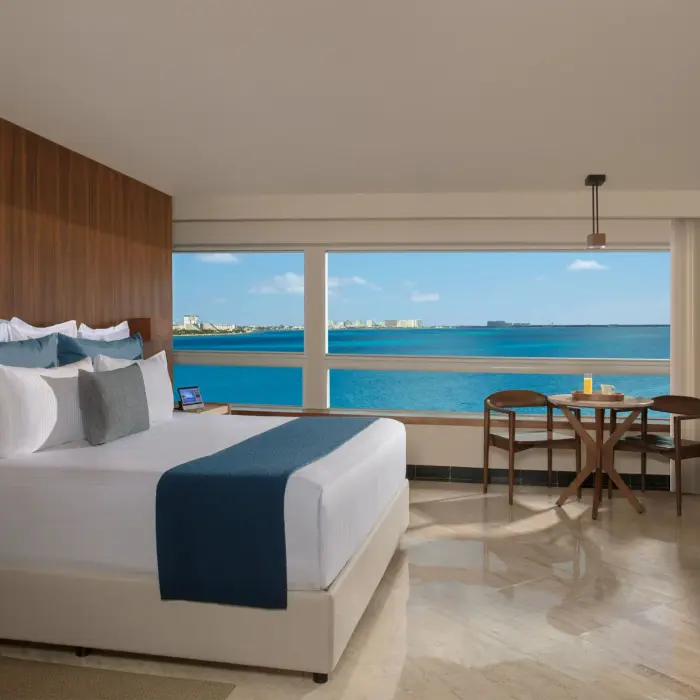 Oceanfront suite with king bed, wood panel accents, work desk, and panoramic views from wide corner windows.