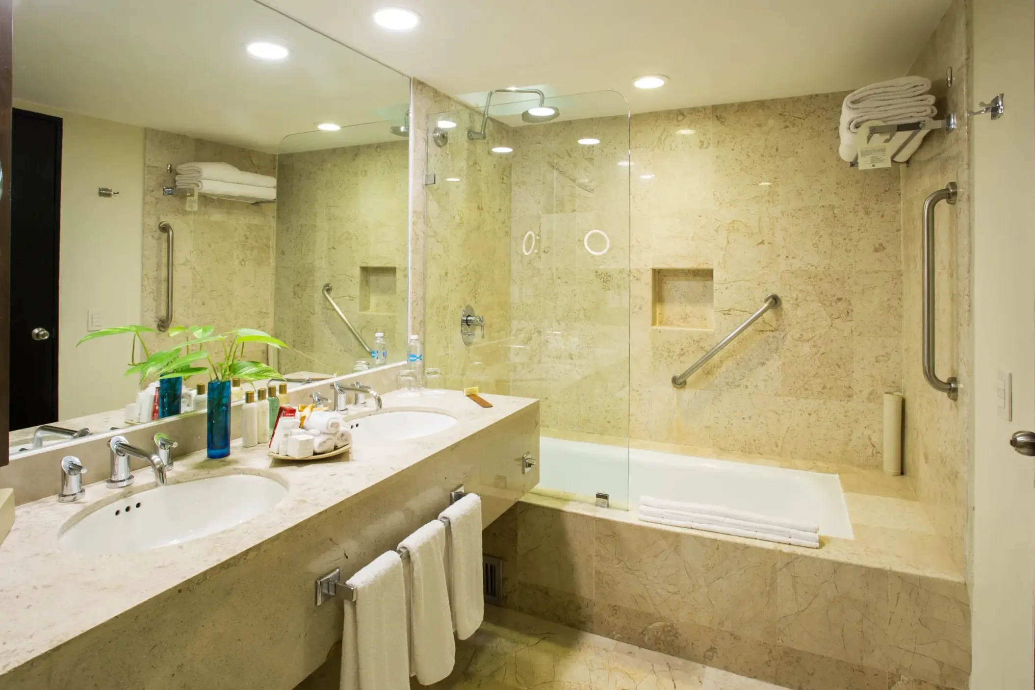 Marble bathroom with glass walk-in shower, tub, dual vanity, and towel racks under soft ceiling lighting.