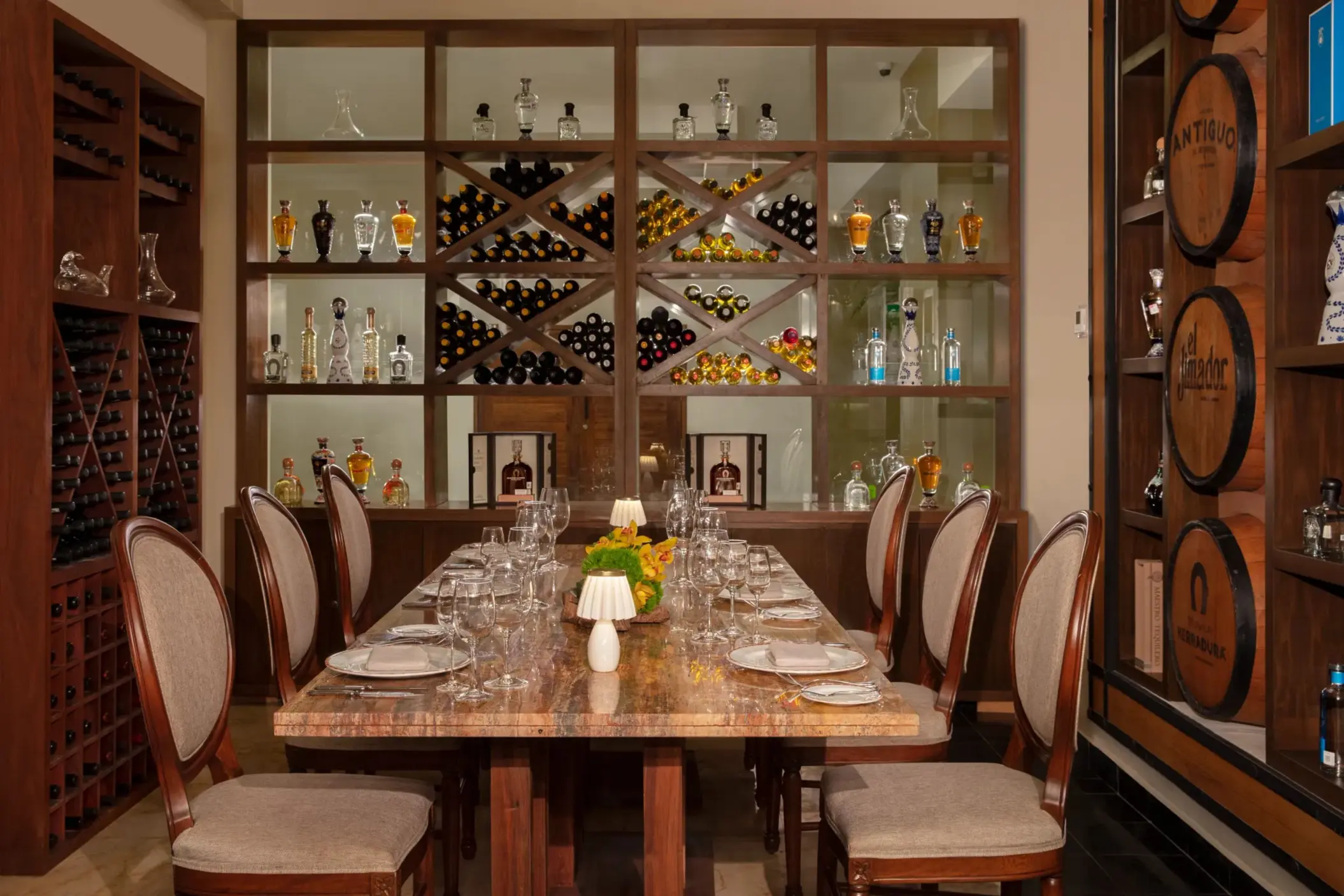 Refined wine cellar dining room with marble table, plush chairs, and backlit shelves of bottles and barrels.