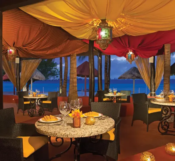 Romantic Olio beachfront dining under colorful canopy with ocean view and candlelit tables.