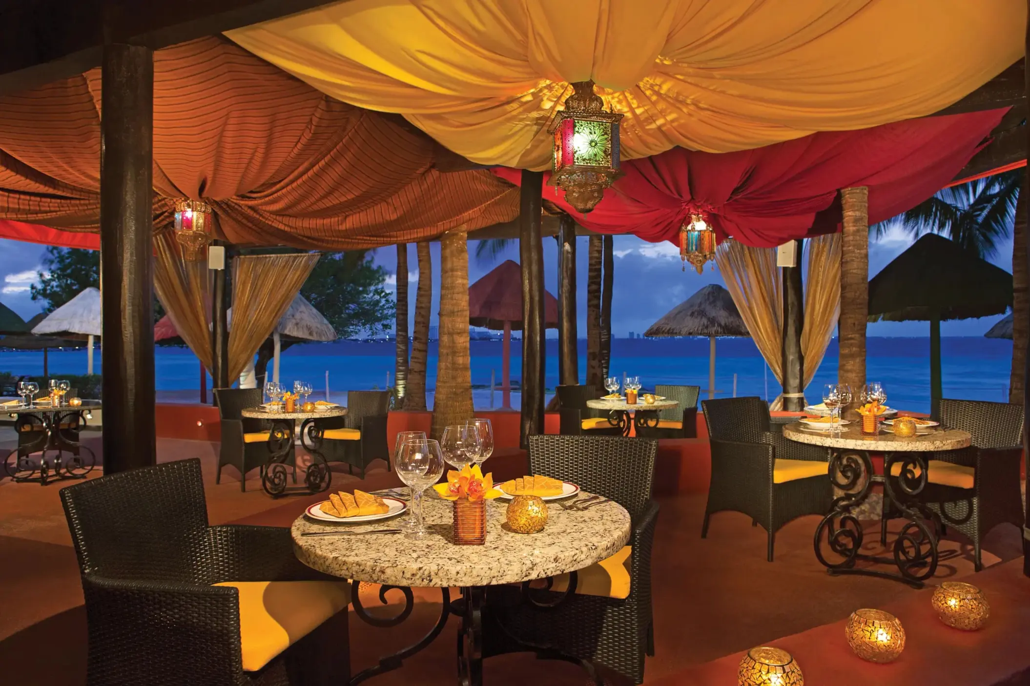 Romantic Olio beachfront dining under colorful canopy with ocean view and candlelit tables.