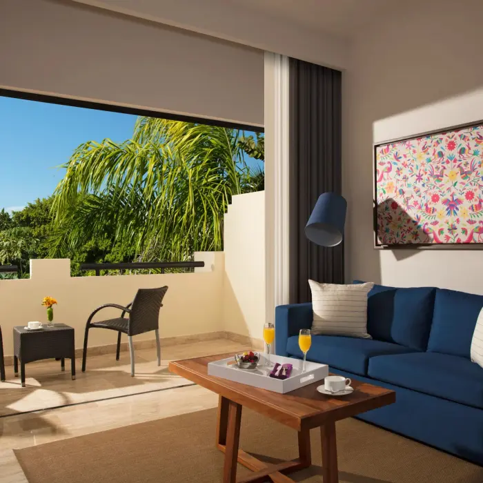 Spacious suite with private balcony, two chairs and table, tropical views, and comfortable seating inside.