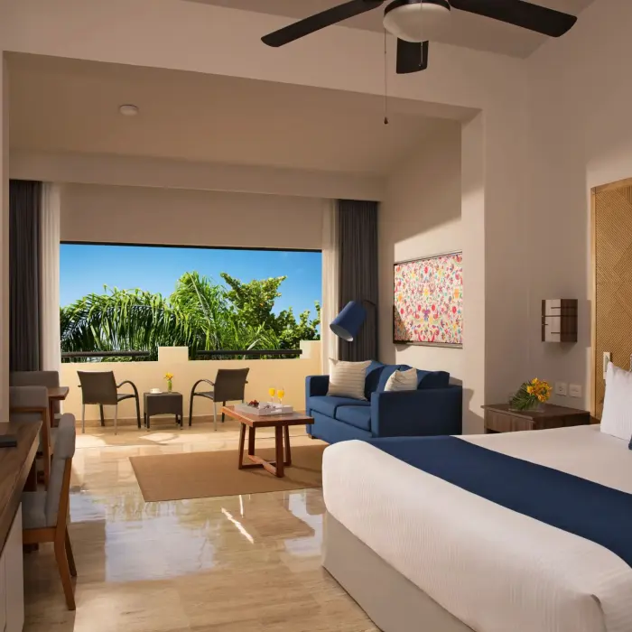 Spacious resort suite with king bed, seating area, and balcony view of tropical gardens and blue skies.