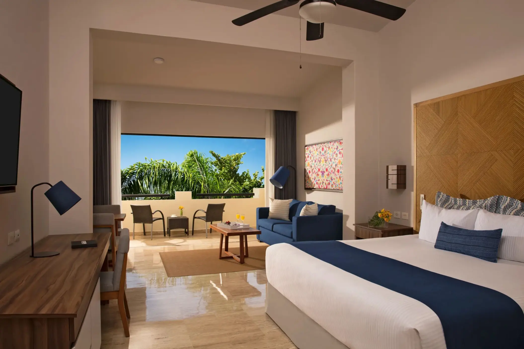 Spacious resort suite with king bed, seating area, and balcony view of tropical gardens and blue skies.