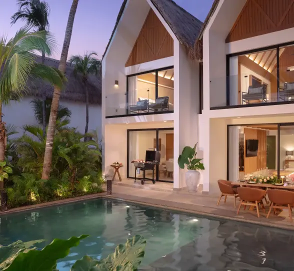 Two-story private villa at dusk with a pool, tropical plants, and a patio dining area with a grill.