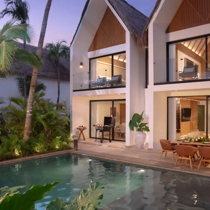 Two-story private villa at dusk with a pool, tropical plants, and a patio dining area with a grill.
