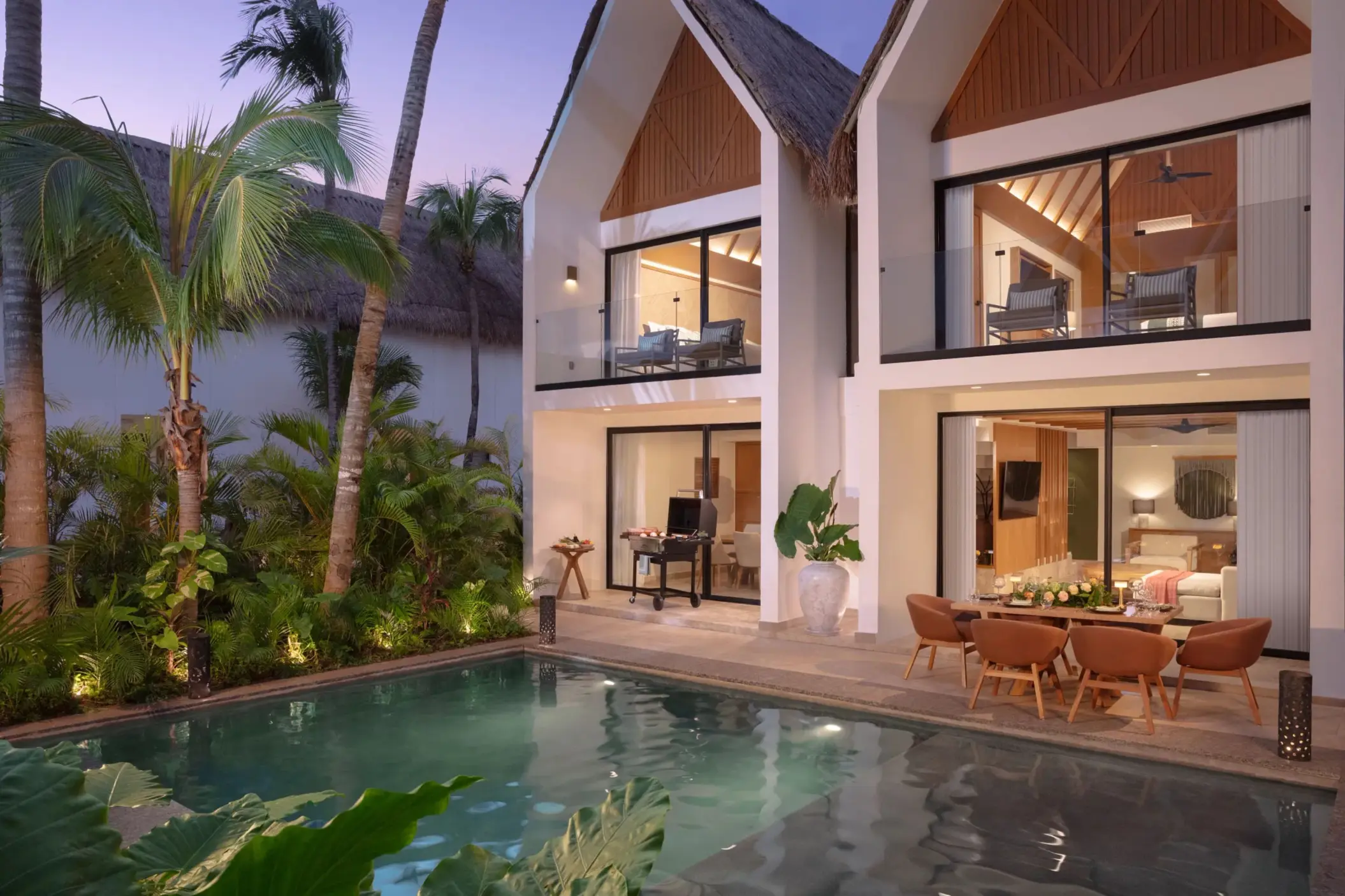 Two-story private villa at dusk with a pool, tropical plants, and a patio dining area with a grill.