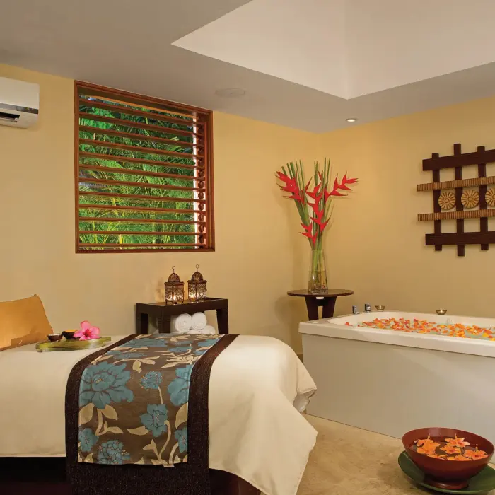 Tranquil spa treatment room with massage bed, floral accents, and a deep soaking tub filled with petals.