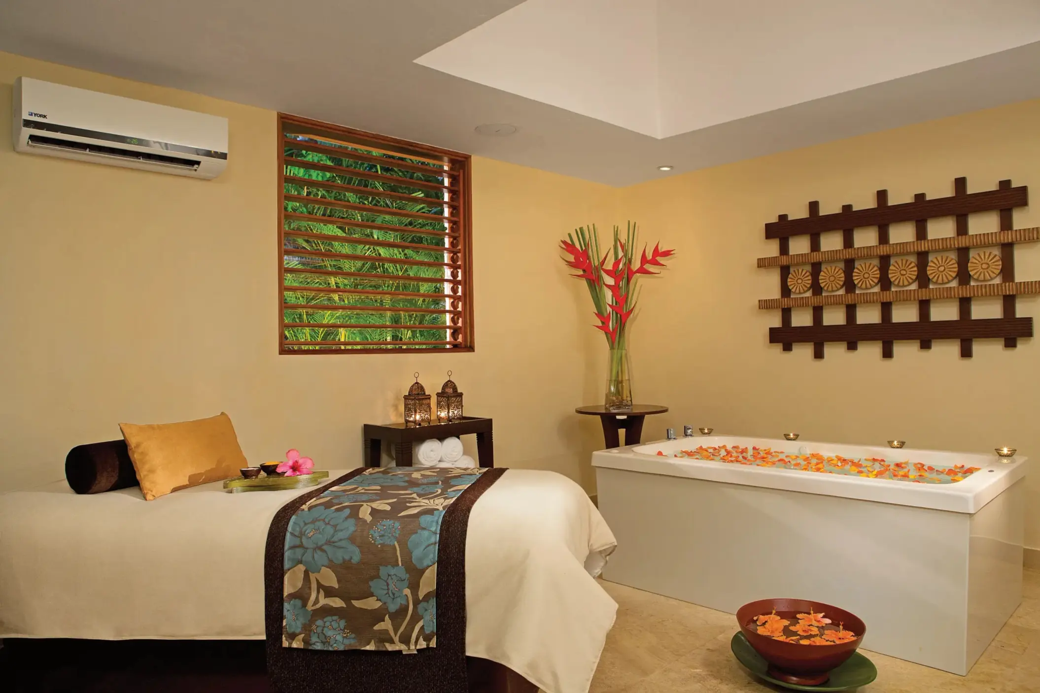 Tranquil spa treatment room with massage bed, floral accents, and a deep soaking tub filled with petals.