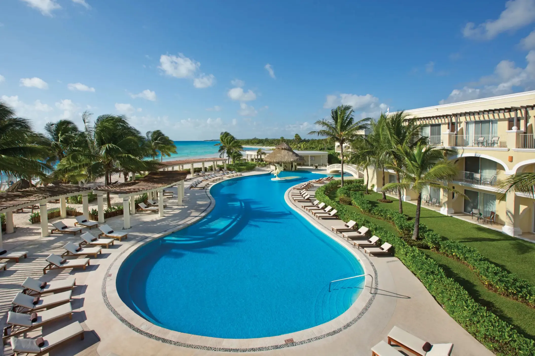 Large freeform pool at Dreams Tulum surrounded by sun loungers, palm trees, and beachfront rooms with ocean views.