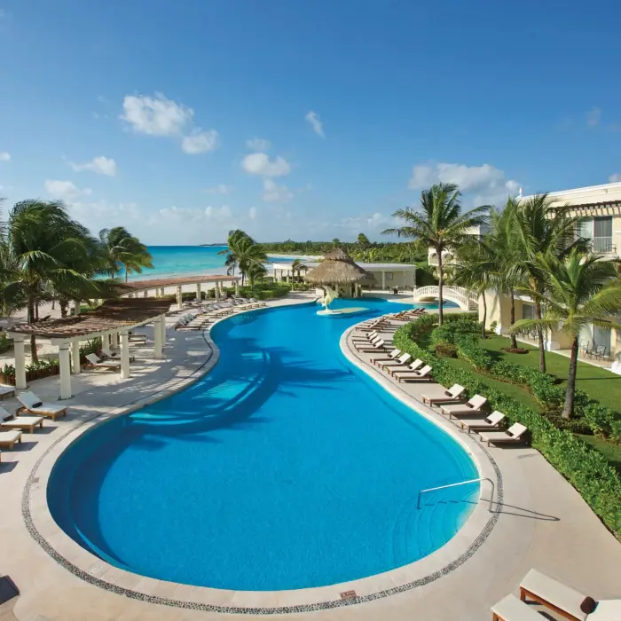 Large freeform pool at Dreams Tulum surrounded by sun loungers, palm trees, and beachfront rooms with ocean views.