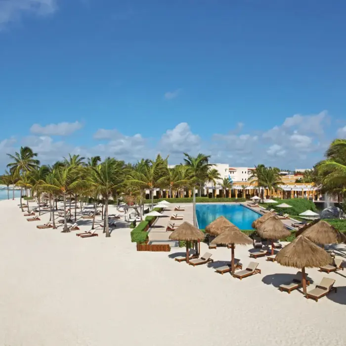 Aerial view of Dreams Tulum beachfront with palapas, loungers, palm trees, and a central pool under clear blue skies.