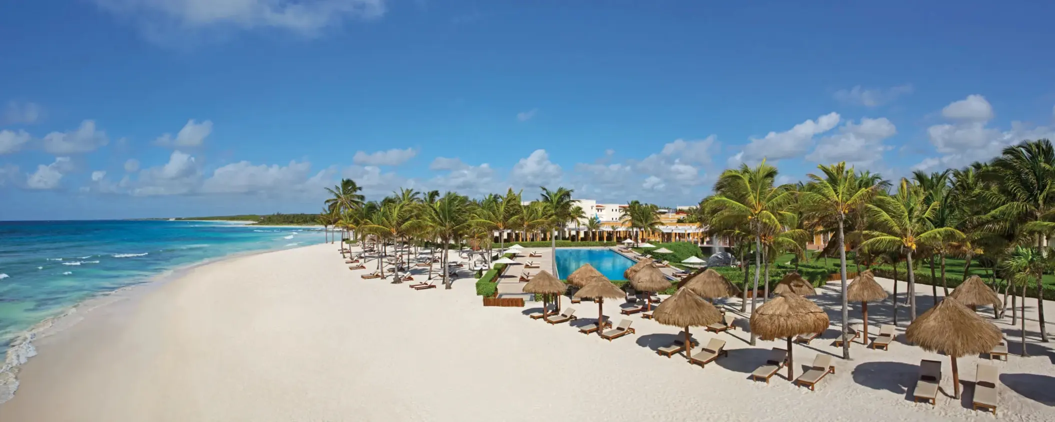 Aerial view of Dreams Tulum beachfront with palapas, loungers, palm trees, and a central pool under clear blue skies.
