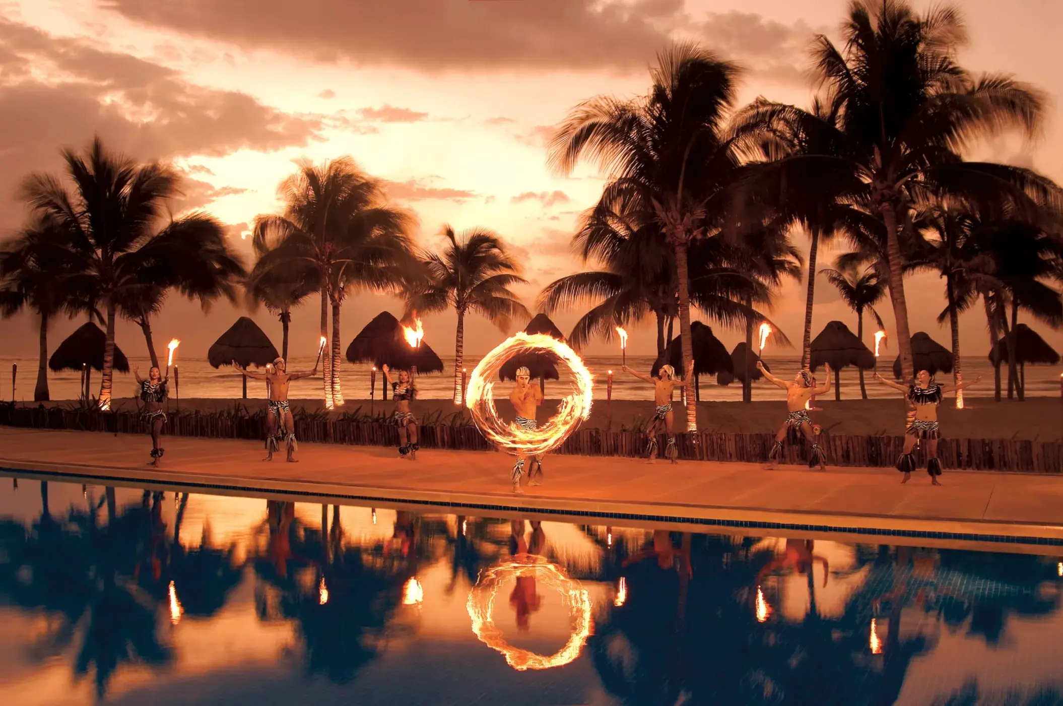 Fire dancers perform at sunset near the beach with palm trees silhouetted against a glowing sky.