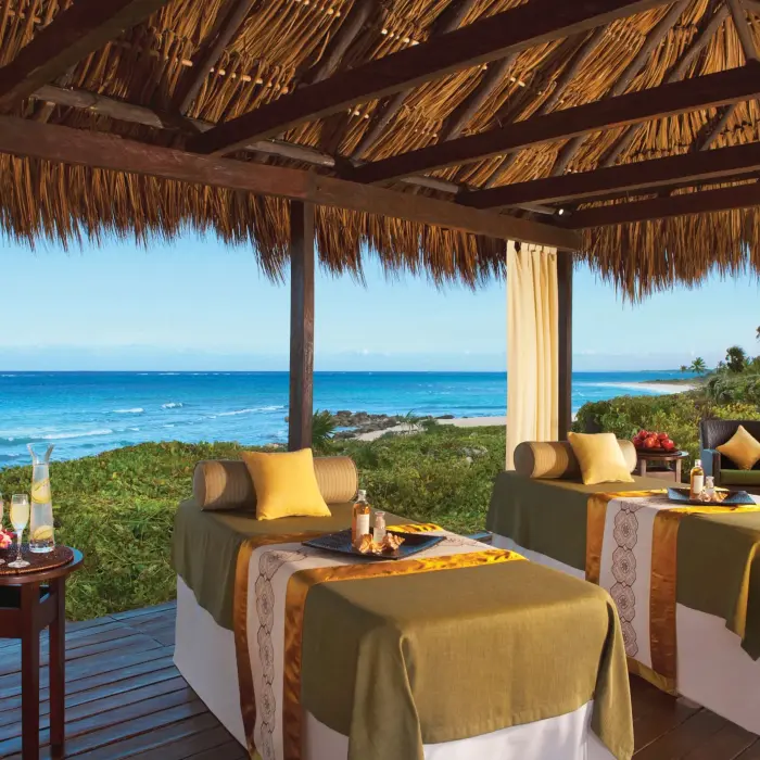 Oceanfront spa cabin with two massage tables under a palapa roof, overlooking the turquoise sea.