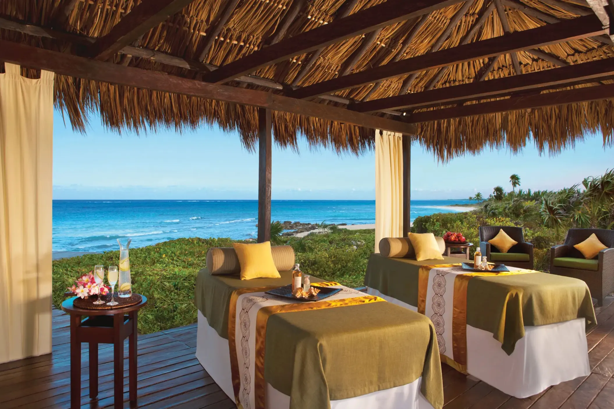 Oceanfront spa cabin with two massage tables under a palapa roof, overlooking the turquoise sea.