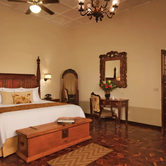 Elegant colonial-style suite with king bed, vintage wood furnishings, and separate sitting area with warm lighting.